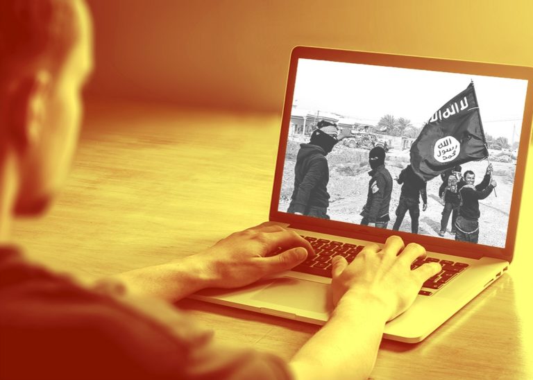 A man sitting at a desk using a laptop. On the laptop screen, a group of ISIS members, some with their faces covered, hoist the ISIS flag.